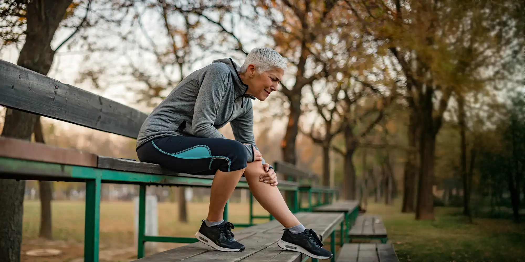 Physical Medicine Columbus GA Person With Knee Pain On Park Bleachers Middle Aged Woman Sitting On Park Bleachers Clutching Her Knee In Pain